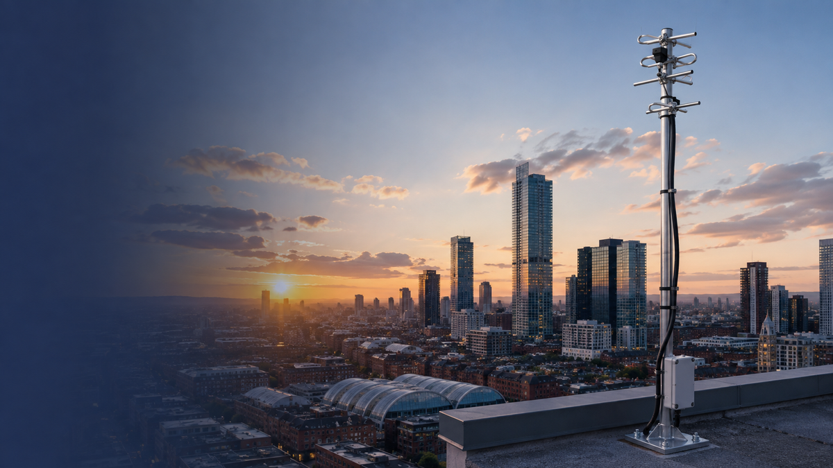 A tall metal antenna mast mounted on a rooftop overlooking a city skyline at sunset, with skyscrapers, low-rise buildings, and a blue-to-orange sky.
